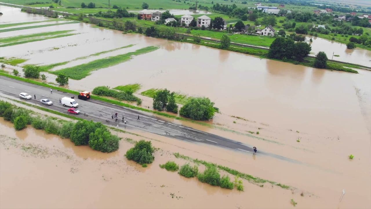 Aerial view of a road disappearing into brown floodwater, with cars and people stopped at the edge