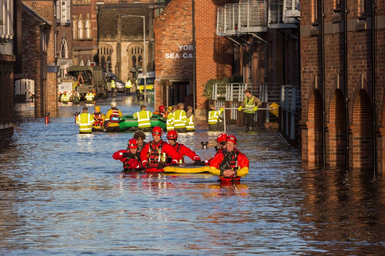 Rescue teams in drysuits and hi-vis wade through a flooded street with inflatable boats